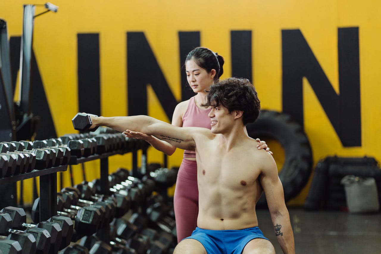 Young man lifting weights with a woman trainer assisting in a gym setting.